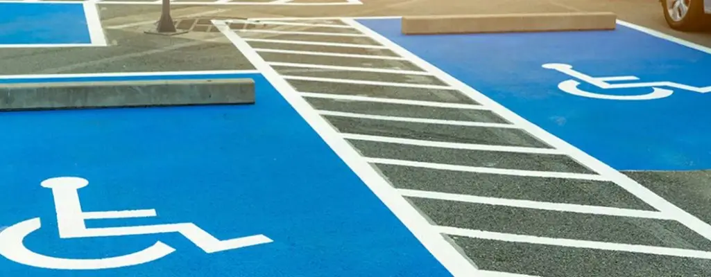 Obrazek wyróżniający do artykułu - miejsce parkingowe dla niepełnosprawnych. Obraz przedstawia parking z miejscem przeznaczonym dla osób niepełnosprawnych, oznaczonym niebieskim kolorem i symbolem wózka inwalidzkiego. Na miejscu tym zaparkowany jest czarny samochód, a przez środek miejsca parkingowego przebiega latarnia uliczna. Obok, po prawej stronie, zaparkowany jest kolejny czarny samochód. W tle widać budynki mieszkalne oraz ogrodzenie z żywopłotem. Całość otoczenia wskazuje na miejską okolicę z blokami mieszkalnymi i miejscami parkingowymi.
