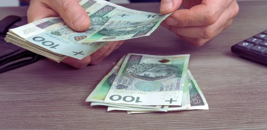 Obrazek wyróżniający do artykułu - świadczenia dla niepełnosprawnych. Na obrazie widoczne są ręce osoby liczącej polskie banknoty o nominale 100 złotych. Banknoty są starannie układane na stole, a obok znajduje się kalkulator, co sugeruje wykonywanie obliczeń finansowych. Obraz symbolizuje zarządzanie pieniędzmi, wypłaty lub świadczenia finansowe.