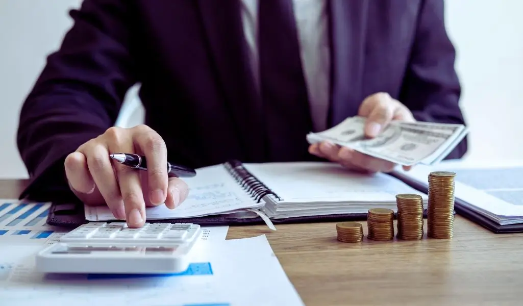 Obrazek do artykułu - warunki otrzymania dofinansowania. Na obrazie widzimy osobę w formalnym stroju, skupioną na obliczeniach finansowych. Na biurku leży kalkulator, stos dokumentów oraz równiutko ułożone stosy monet, symbolizujące uporządkowane zarządzanie środkami finansowymi. W ręku osoba trzyma plik banknotów, co wywołuje skojarzenia z kontrolą nad budżetem i analizą warunków otrzymania dofinansowania. Obraz emanuje profesjonalizmem i precyzją, podkreślając znaczenie dokładności w procesie ubiegania się o środki.