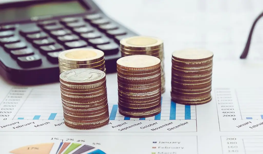 Na obrazie widzimy stosy monet ułożone w równych kolumnach, które symbolizują stabilność finansową oraz gromadzenie środków. W tle znajduje się kalkulator oraz wykresy na papierze, co sugeruje analizę finansową i strategiczne planowanie. Kolorystyka obrazu utrzymana jest w jasnych, stonowanych tonach, co wprowadza uczucie profesjonalizmu i precyzji. Motyw ten doskonale oddaje temat związany z zarządzaniem finansami, takimi jak dofinansowanie wynagrodzeń PFRON w 2025, podkreślając znaczenie skutecznej alokacji środków na realizację programów wsparcia.