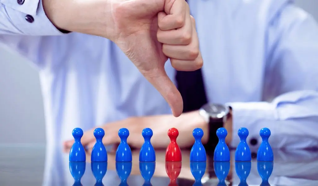 Na obrazku widzimy rząd niebieskich pionków oraz jeden czerwony pionek pośrodku, który wyróżnia się kolorem. Nad czerwonym pionkiem unosi się ręka w koszuli, wykonująca gest "kciuk w dół", symbolizujący dezaprobatę lub odrzucenie. Obraz przywołuje silne skojarzenia z izolacją, odrzuceniem i brakiem akceptacji. Przedstawia doświadczenie jednostki, która różni się od reszty i jest negatywnie oceniana, mimo że nie ma ku temu widocznych powodów. Zdjęcie świetnie oddaje emocje towarzyszące wykluczeniu i nierównemu traktowaniu w miejscu pracy, idealnie ilustrując temat, jakim jest dyskryminacja pośrednia w pracy.