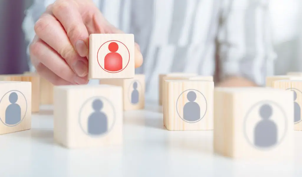 Na obrazku widać dłoń osoby, która sięga po drewniany klocek z czerwonym symbolem człowieka, wyróżniającym się na tle innych klocków z szarymi sylwetkami. Klocek ten symbolizuje wybór, decyzję, a także zauważenie kogoś wyjątkowego wśród wielu. Całość sugeruje proces rekrutacji lub selekcji kandydatów. Obraz wywołuje skojarzenia z poszukiwaniem talentów, wyróżnianiem potencjału oraz tworzeniem miejsca dla osób, które często są pomijane. Może symbolizować także próbę zobaczenia wartości tam, gdzie inni jej nie dostrzegli. Symboliczny obraz ilustrujący temat gdzie szukać pracowników niepełnosprawnych, wyrażający decyzję, uważność i docenienie różnorodności.