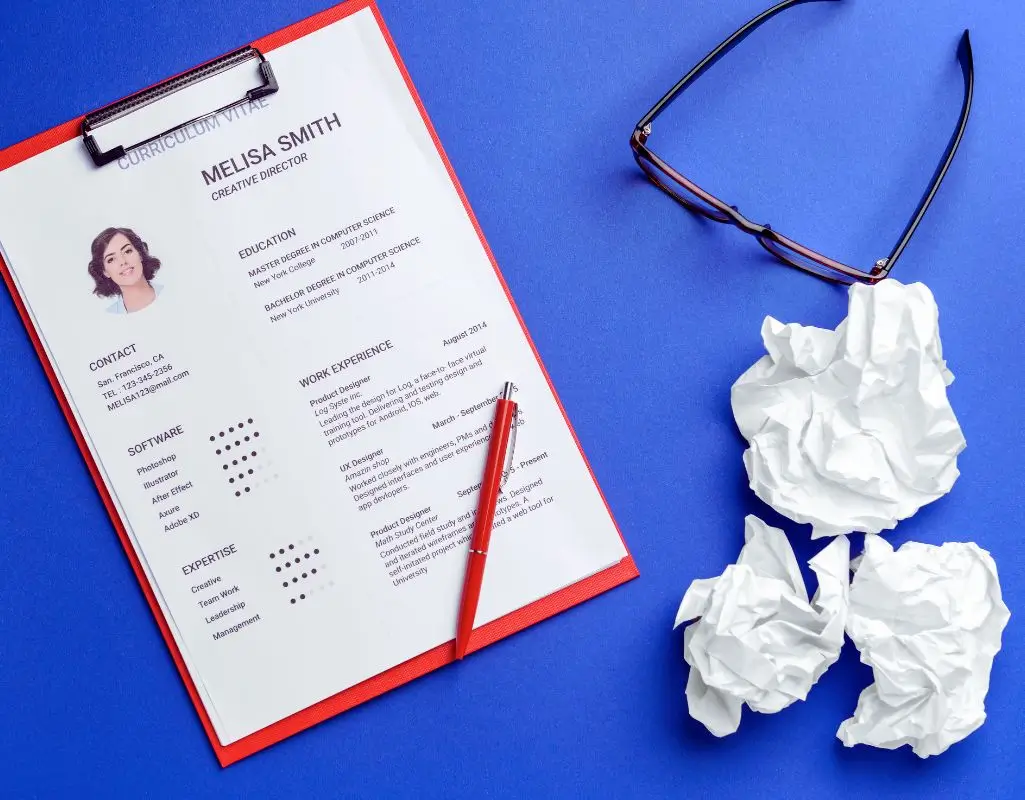 Na zdjęciu widoczny jest starannie przygotowany dokument zatytułowany „Curriculum Vitae” – przykładowy życiorys. CV zostało umieszczone na czerwonym clipboardzie i prezentuje dane kontaktowe, wykształcenie, doświadczenie zawodowe, znajomość programów graficznych oraz umiejętności miękkie. Obok znajdują się trzy zgniecione kartki papieru – symbol nieudanych prób lub wcześniejszych wersji, które nie spełniły oczekiwań. Po prawej stronie leżą również okulary i czerwony długopis, co dodaje scenie osobistego, codziennego charakteru. Całość utrzymana jest w tonacji niebiesko-czerwonej, co przyciąga wzrok i nadaje dynamiki. Obrazek doskonale oddaje atmosferę koncentracji, starań i poprawek, które towarzyszą tworzeniu idealnego CV. Może być inspiracją dla osób szukających podpowiedzi, jak napisać CV wzór – łącząc profesjonalizm z osobistym zaangażowaniem.