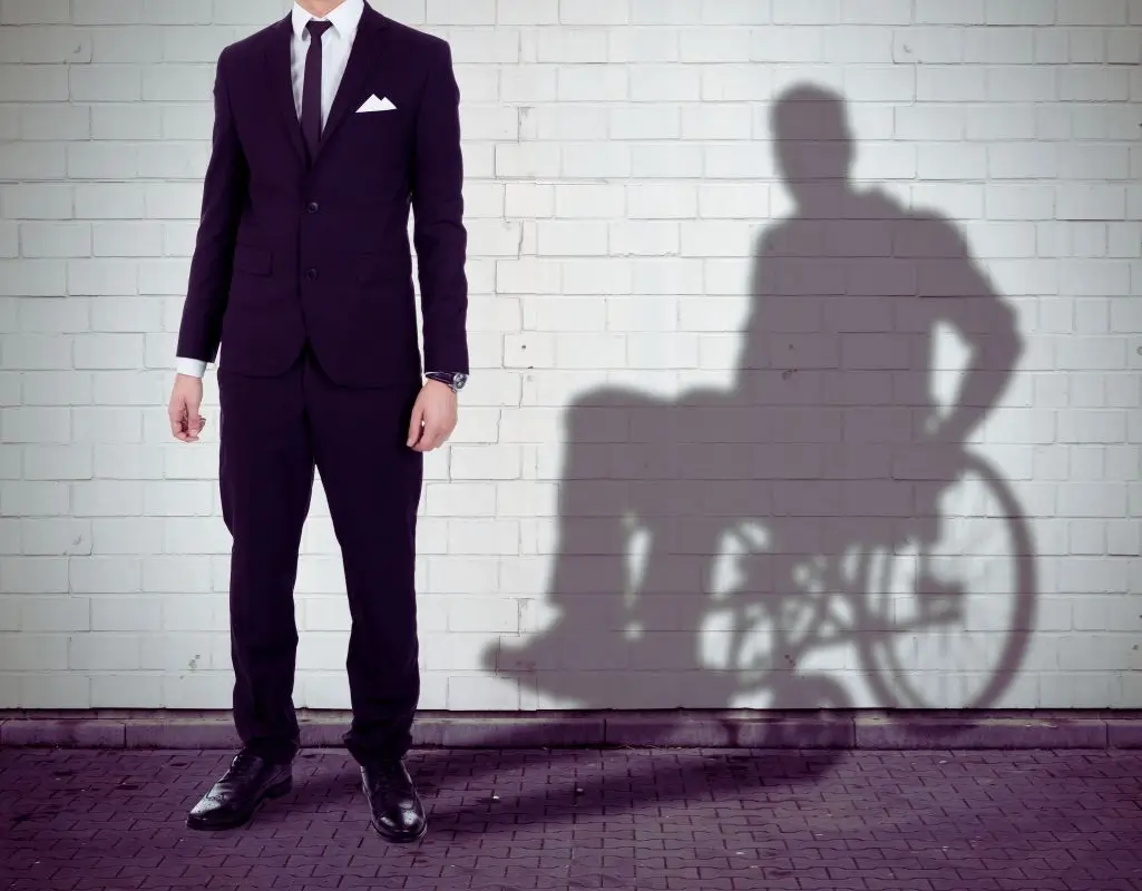 Mężczyzna w eleganckim garniturze stoi przed jasną ścianą z cegieł, jednak jego cień pokazuje postać siedzącą na wózku inwalidzkim. Obraz symbolicznie ukazuje niewidoczne bariery i doświadczenia, z jakimi może mierzyć się osoba z niepełnosprawnością, mimo że na zewnątrz wydaje się „pełnosprawna”. To poruszająca wizualizacja tematu, jakim jest dyskryminacja osób niepełnosprawnych w pracy – sugerująca, że pozory mogą mylić, a prawdziwe wyzwania bywają niewidoczne dla innych. Zdjęcie budzi refleksję nad równością, tożsamością i potrzebą empatii w środowisku zawodowym.